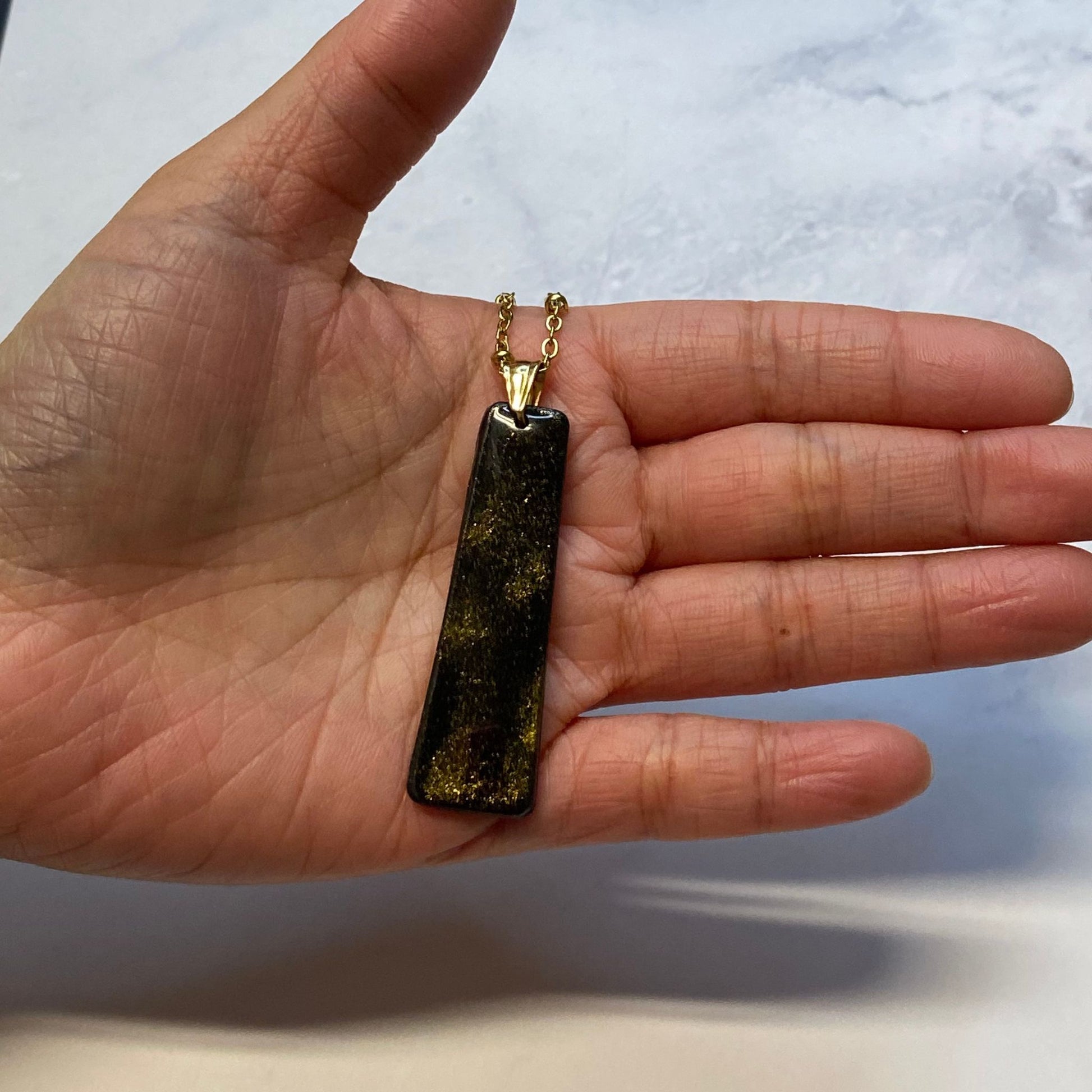 Collana pendente con ciondolo rettangolare allungato in porcellana fredda, colore nero e oro, con catenina dorata. 
Gioiello artigianale fatto a mano da persone cieche e ipovedenti.  
La collana viene tenuta in mano. 