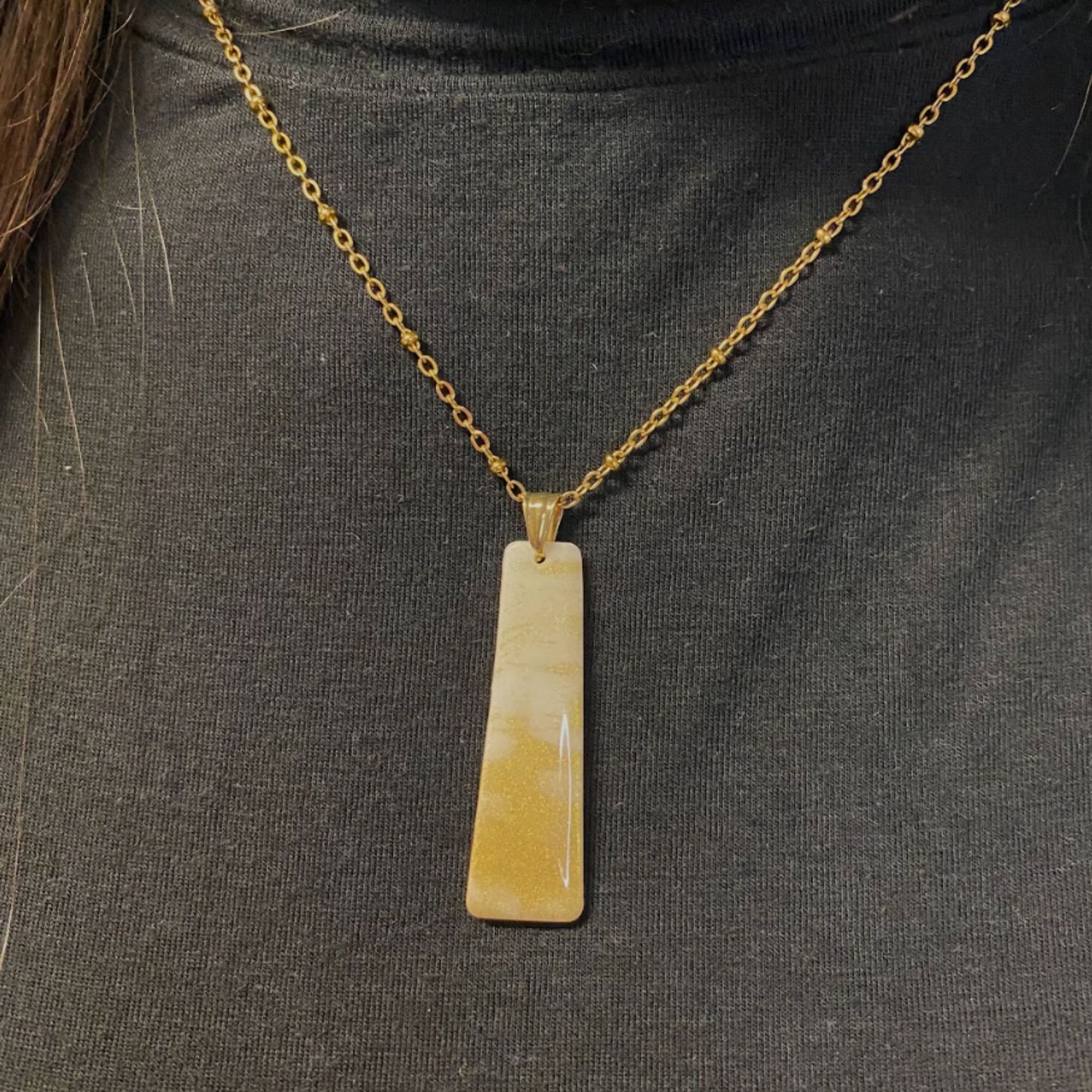 Primo piano della collana indossata su un dolcevita nero. Collana pendente con ciondolo rettangolare allungato in porcellana fredda, colore bianco e oro, effetto marmo, con catenina dorata. Gioiello artigianale fatto a mano da persone cieche e ipovedenti. 