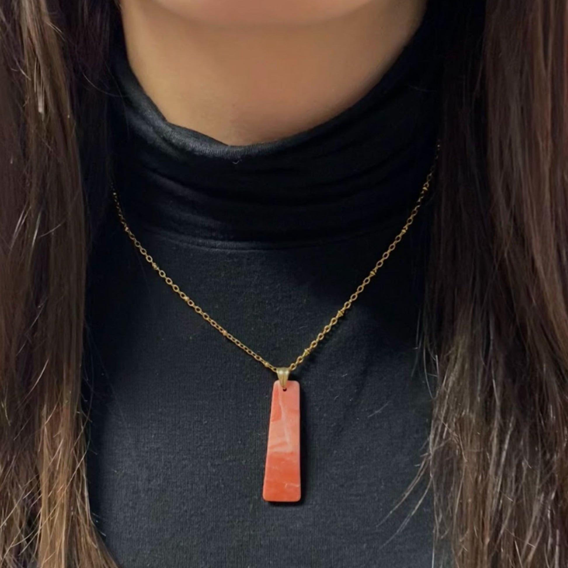 Primo piano della collana indossata su un dolcevita nero. Collana pendente con ciondolo rettangolare allungato in porcellana fredda, colore rosa e bianco, con dettagli oro, effetto marmo, con catenina dorata. Gioiello artigianale fatto a mano da persone cieche e ipovedenti.