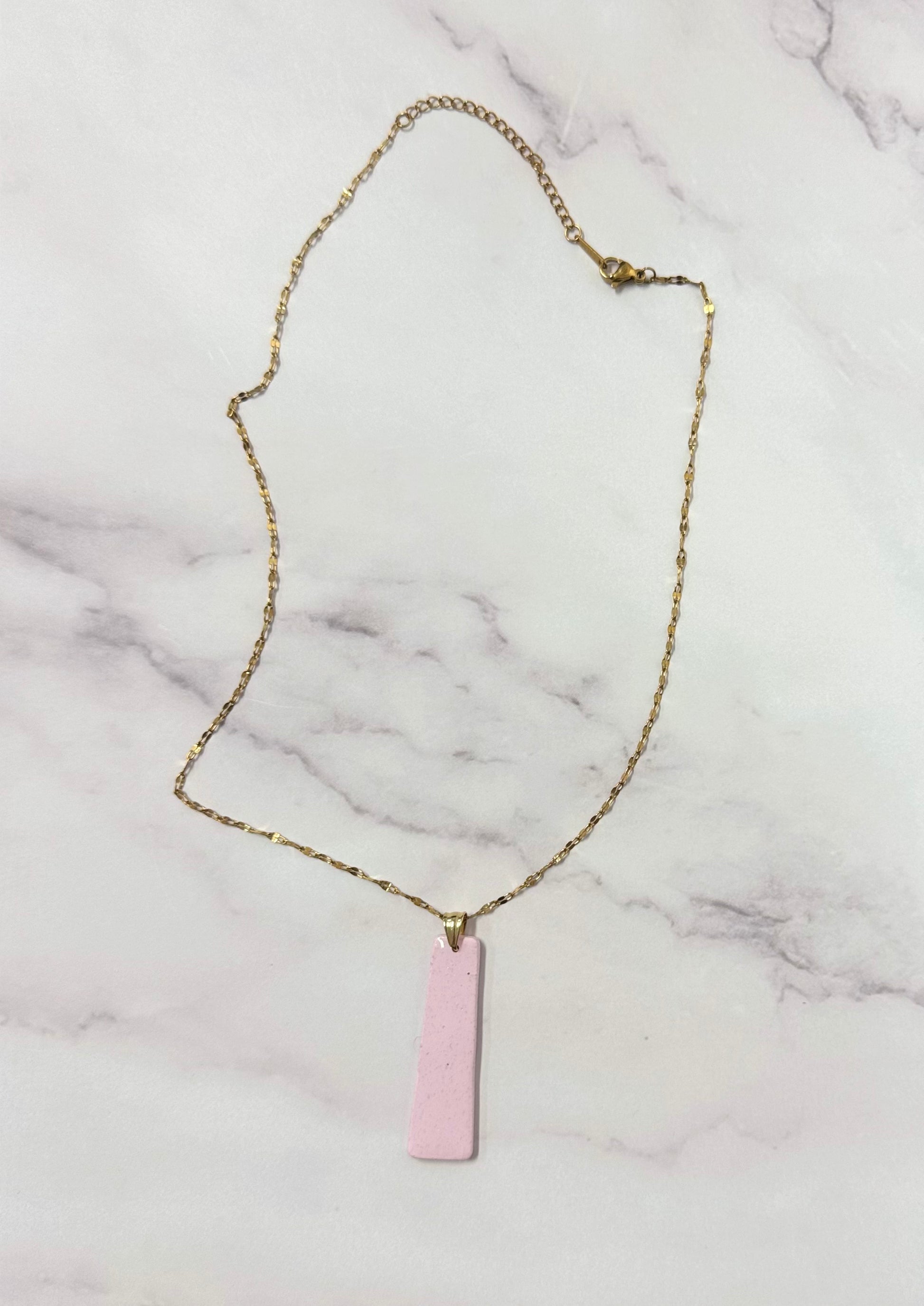 Collana dorata con ciondolo a forma di rettangolo, rosa, in porcellana fredda, adagiata su fondo marmoreo. Creata da artigiani ciechi e ipovedenti.