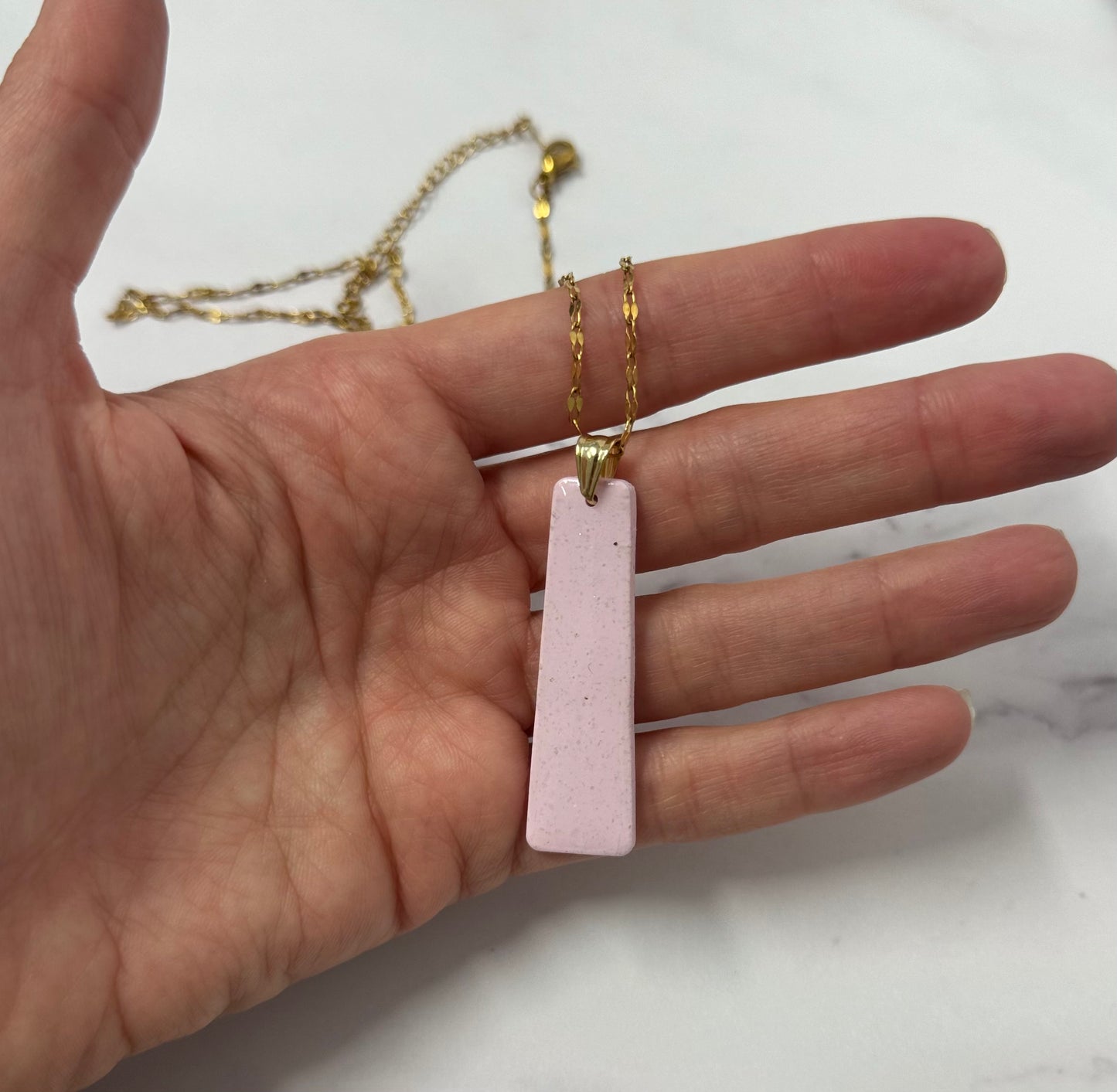 Collana dorata con ciondolo a forma di rettangolo rosa in porcellana fredda, sul palmo di una mano. Gioiello realizzato da artigiani ciechi e ipovedenti.
