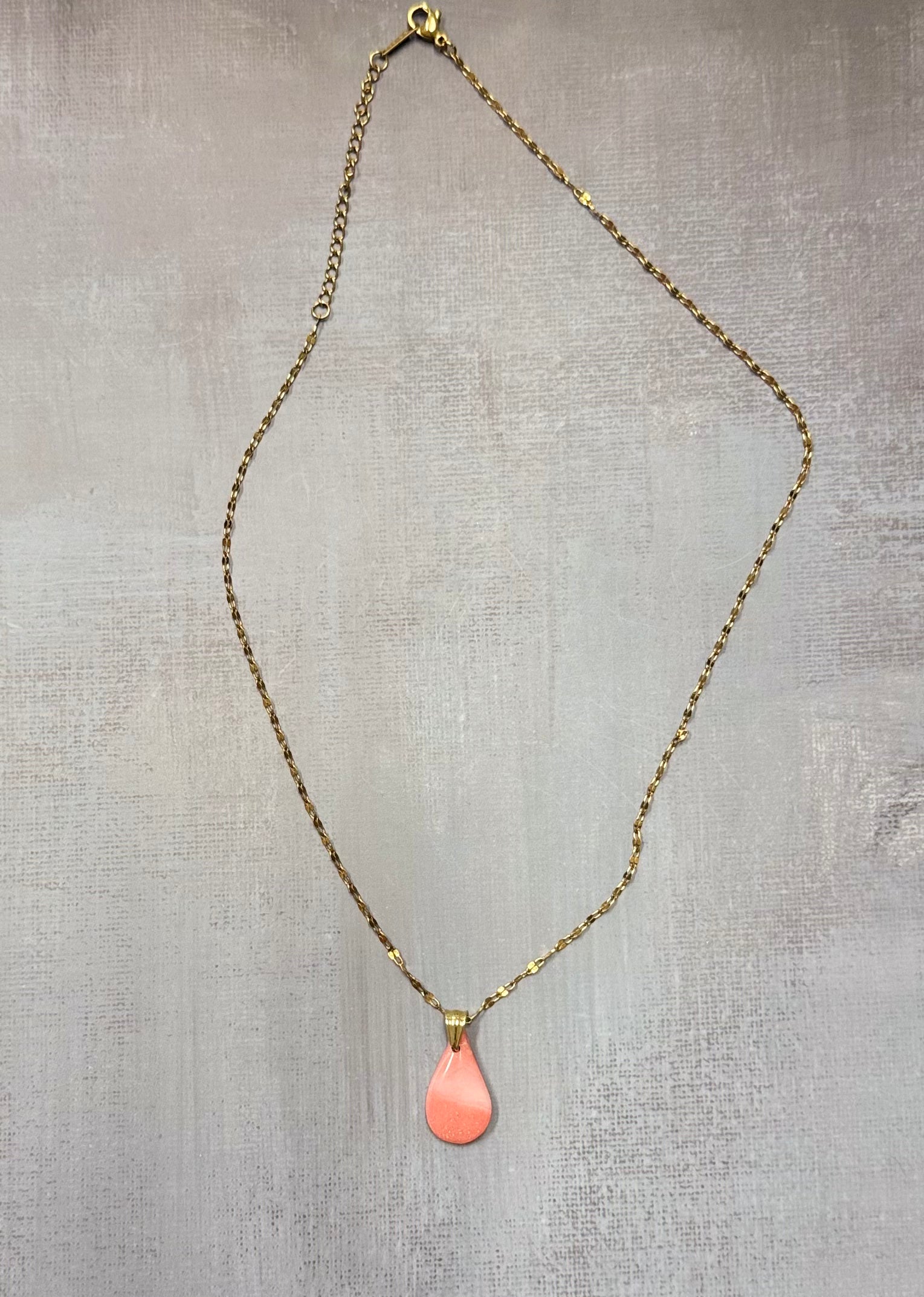 Collana dorata con ciondolo a goccia rosa corallo in porcellana fredda, adagiata su fondo grigio. Creata da artigiani ciechi e ipovedenti.
