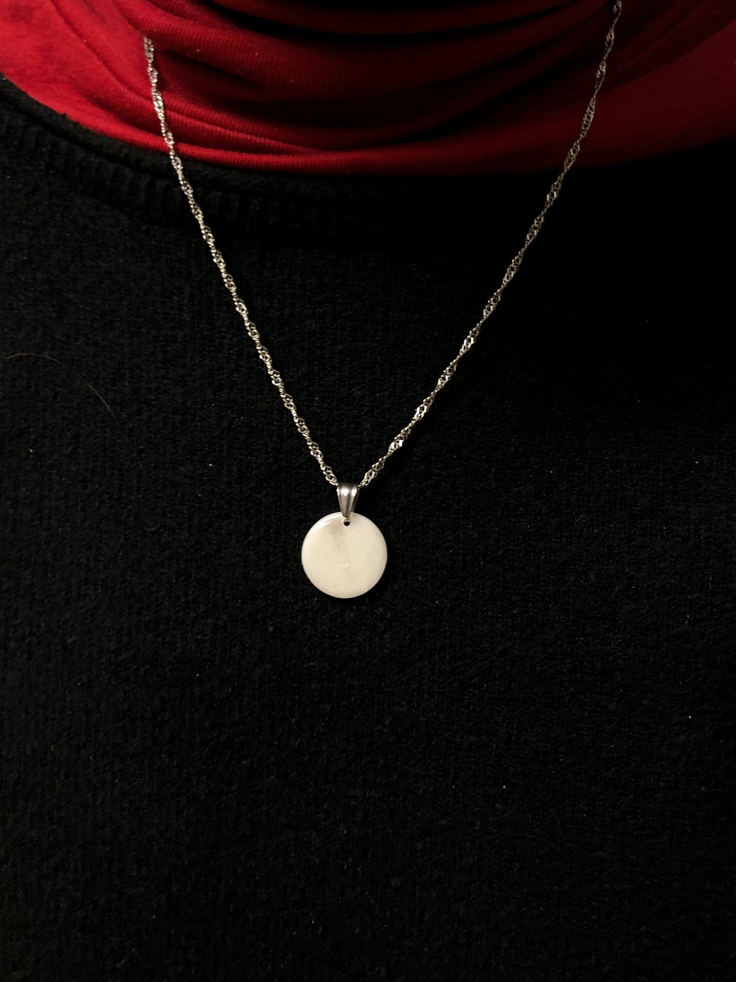 Primo piano della collana indossata su una maglietta nera. Collana pendente con ciondolo a forma di cerchio in porcellana fredda, colore bianco e argento, con catenina argentata. 
Gioiello artigianale fatto a mano da persone cieche e ipovedenti.