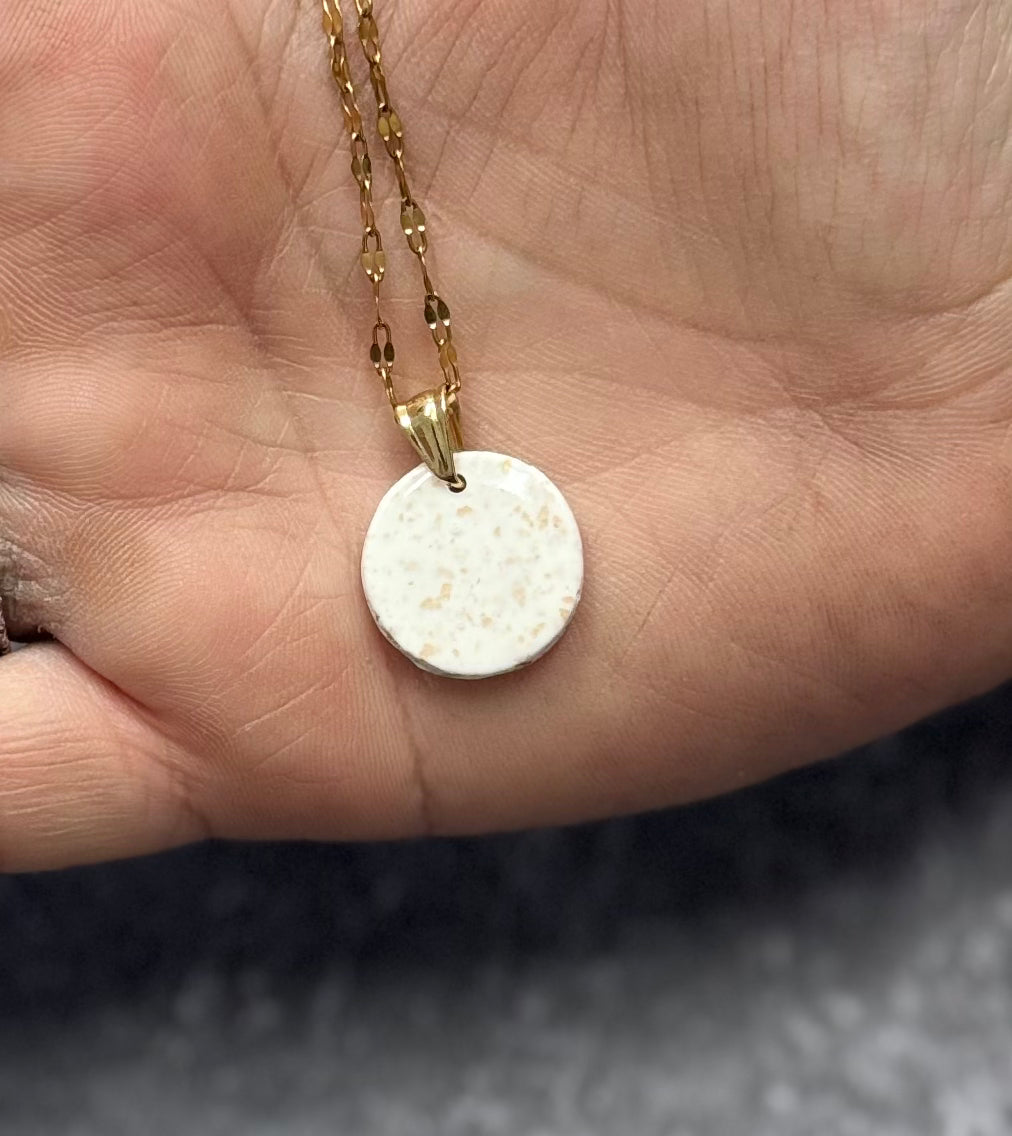 Collana con tondo bianco con pallini dorati in porcellana fredda, tenuta sul palmo di una mano. Gioiello realizzato da artigiani ciechi e ipovedenti