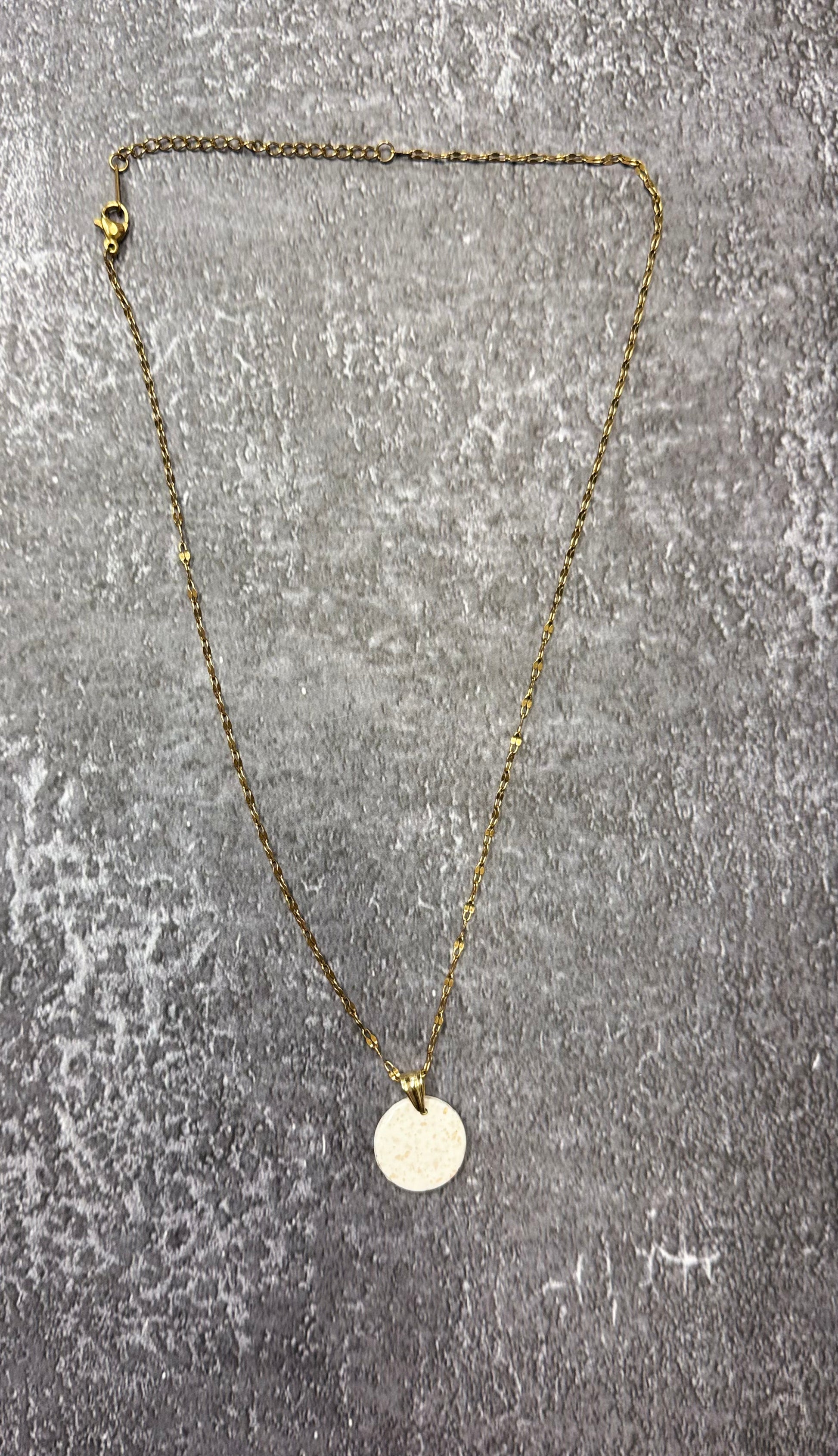 Collana con catenina dorata e pendente tondo bianco con pallini dorati in porcellana fredda e glitter, adagiata su fondo marmoreo. Creata da artigiani ciechi e ipovedenti.