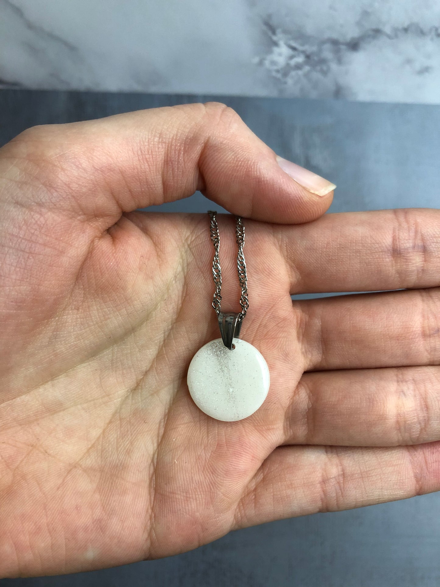 Collana pendente con ciondolo a forma di cerchio in porcellana fredda, colore bianco con dettagli in argento, con catenina argentata. 
Gioiello artigianale fatto a mano da artigiani ciechi e ipovedenti. 
La collana viene tenuta in mano. 