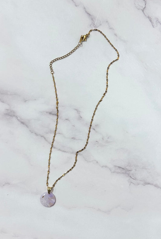 Collana dorata con ciondolo tondo lilla marmorizzato in porcellana fredda e sfumature dorate, adagiata su fondo marmoreo. Creata da artigiani ciechi e ipovedenti