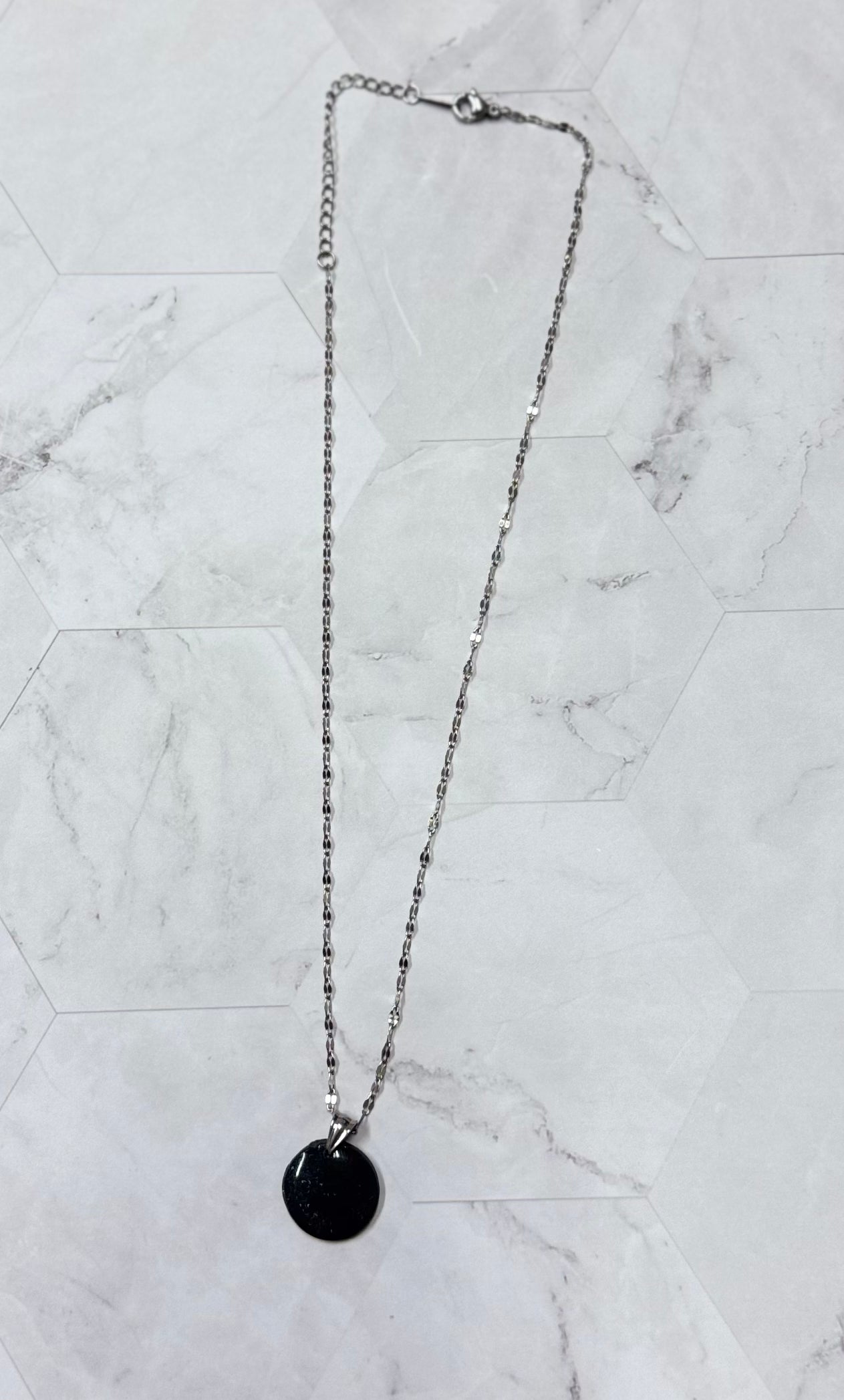 Collana con catenina argentata e pendente tondo nero brillantinato in porcellana fredda, adagiata su fondo marmoreo. Creata da artigiani ciechi e ipovedenti.