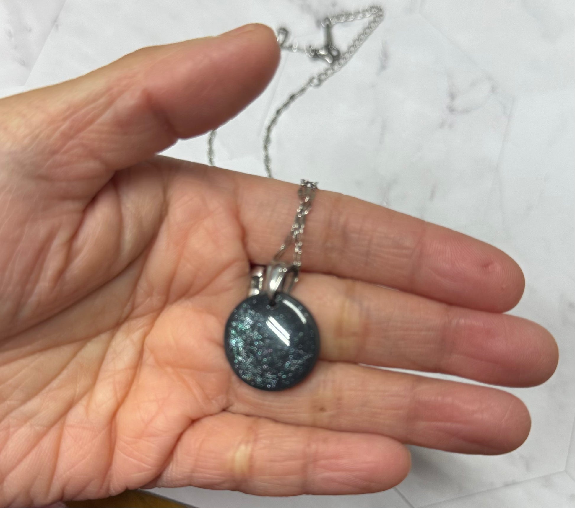 Collana con tondo nero brillantinato in porcellana fredda, tenuta sul palmo di una mano. Gioiello realizzato da artigiani ciechi e ipovedenti