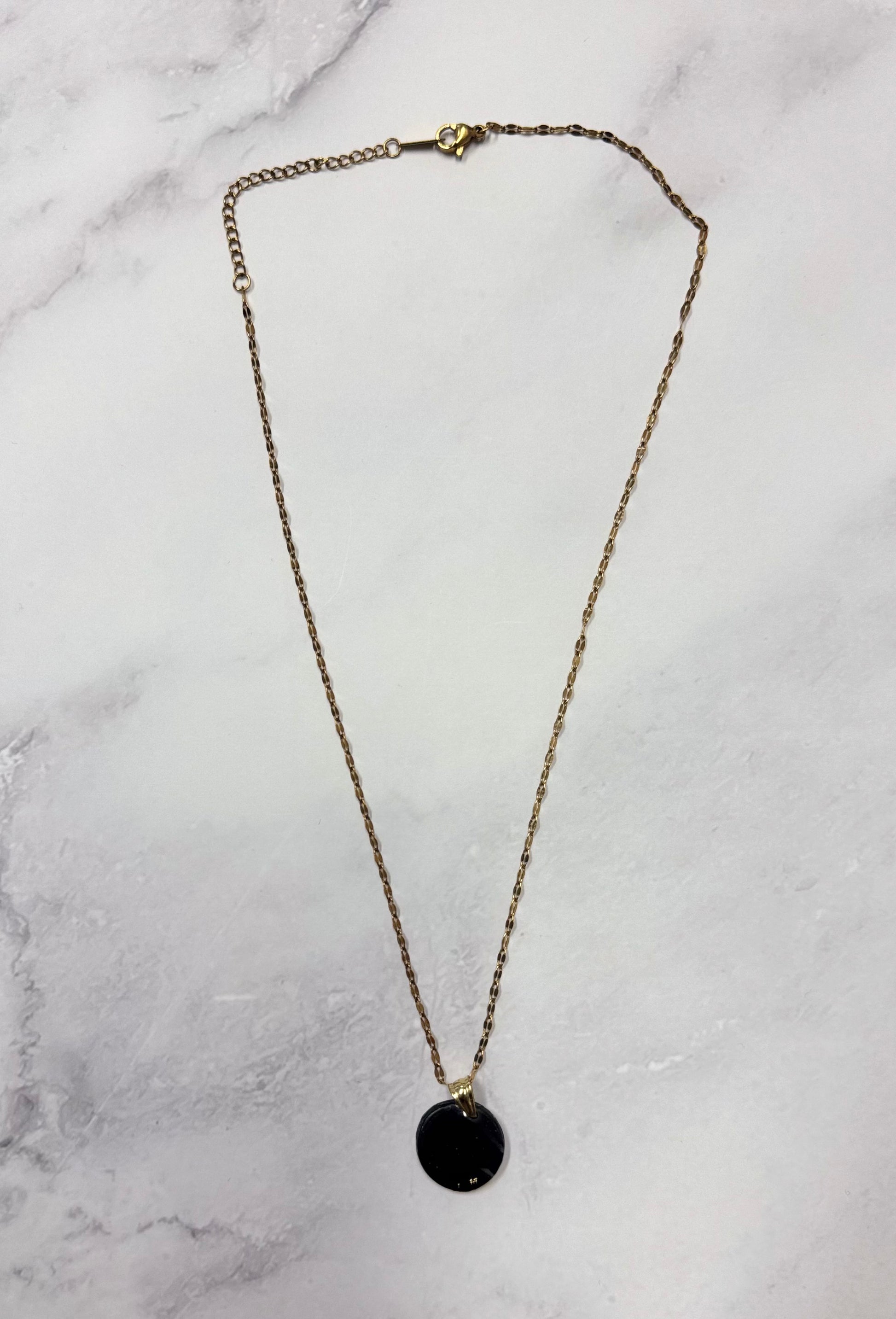 Collana con catenina dorata e pendente tondo nero in porcellana fredda e glitter, adagiata su fondo marmoreo. Creata da artigiani ciechi e ipovedenti.