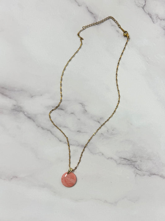 Collana dorata con ciondolo tondo rosso/rosa marmorizzato in porcellana fredda e sfumature dorate, adagiata su fondo marmoreo. Creata da artigiani ciechi e ipovedenti