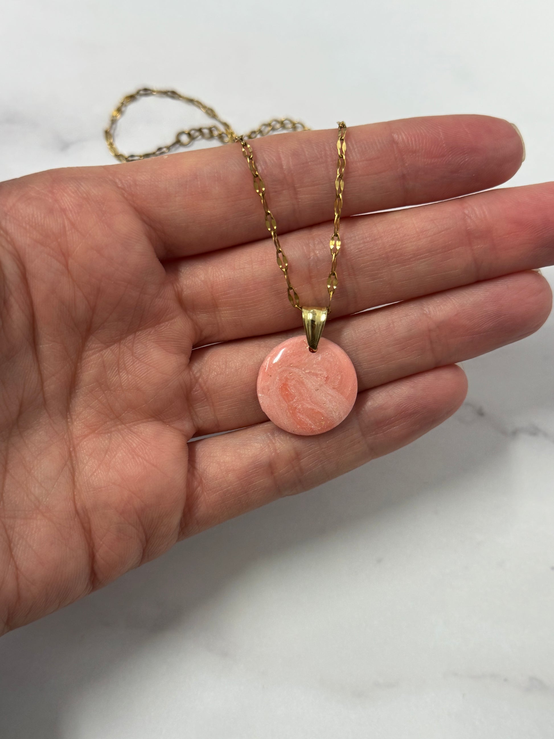 Collana con tondo rosso/rosa marmorizzato in porcellana fredda e leggere sfumature dorate, sul palmo di una mano. Gioiello realizzato da artigiani ciechi e ipovedenti.