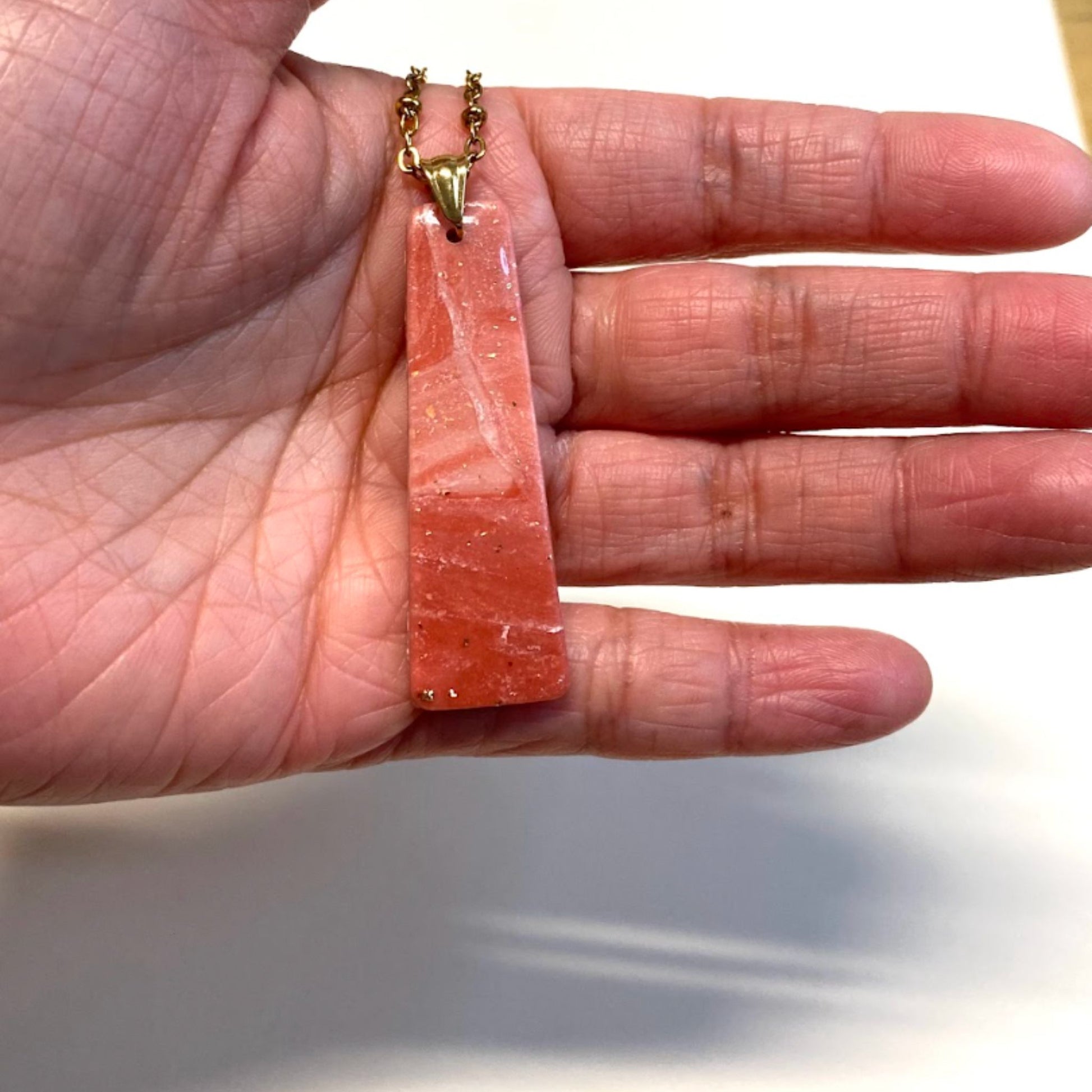 Collana pendente con ciondolo rettangolare allungato in porcellana fredda, colore rosa e bianco, con dettagli oro, effetto marmo, con catenina dorata. Gioiello artigianale fatto a mano da persone cieche e ipovedenti. 
La collana viene tenuta in mano. 
