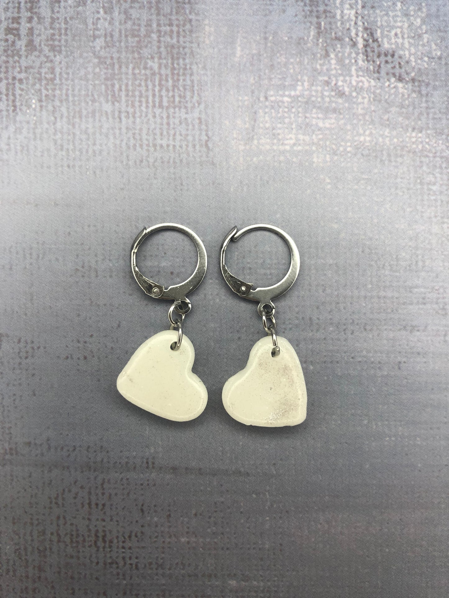 Orecchini pendenti artigianali in porcellana fredda a forma di cuore, colore bianco con sfumature argento, con gancio a cerchio a leva argentato. 
Gioiello realizzato a mano da artigiani ciechi e ipovedenti.
