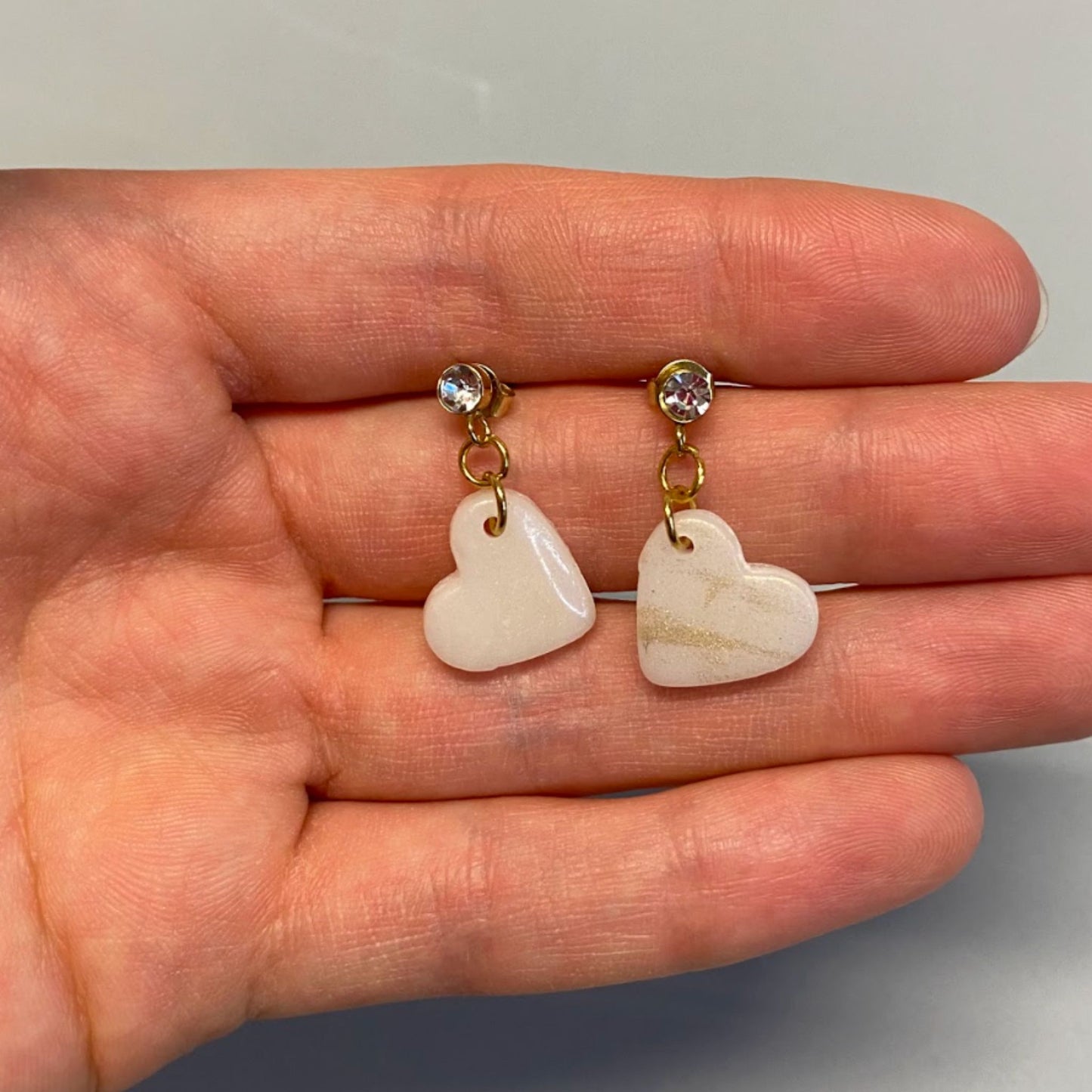Orecchini pendenti handmade in porcellana fredda a forma di cuore, colore bianco con venature dorate.
L'orecchino ha una chiusura a perno dorata, con punto luce in zircone. 
Fatti a mano da artigiani ciechi e ipovedenti.
Gli orecchini sono tenuti in mano. 
