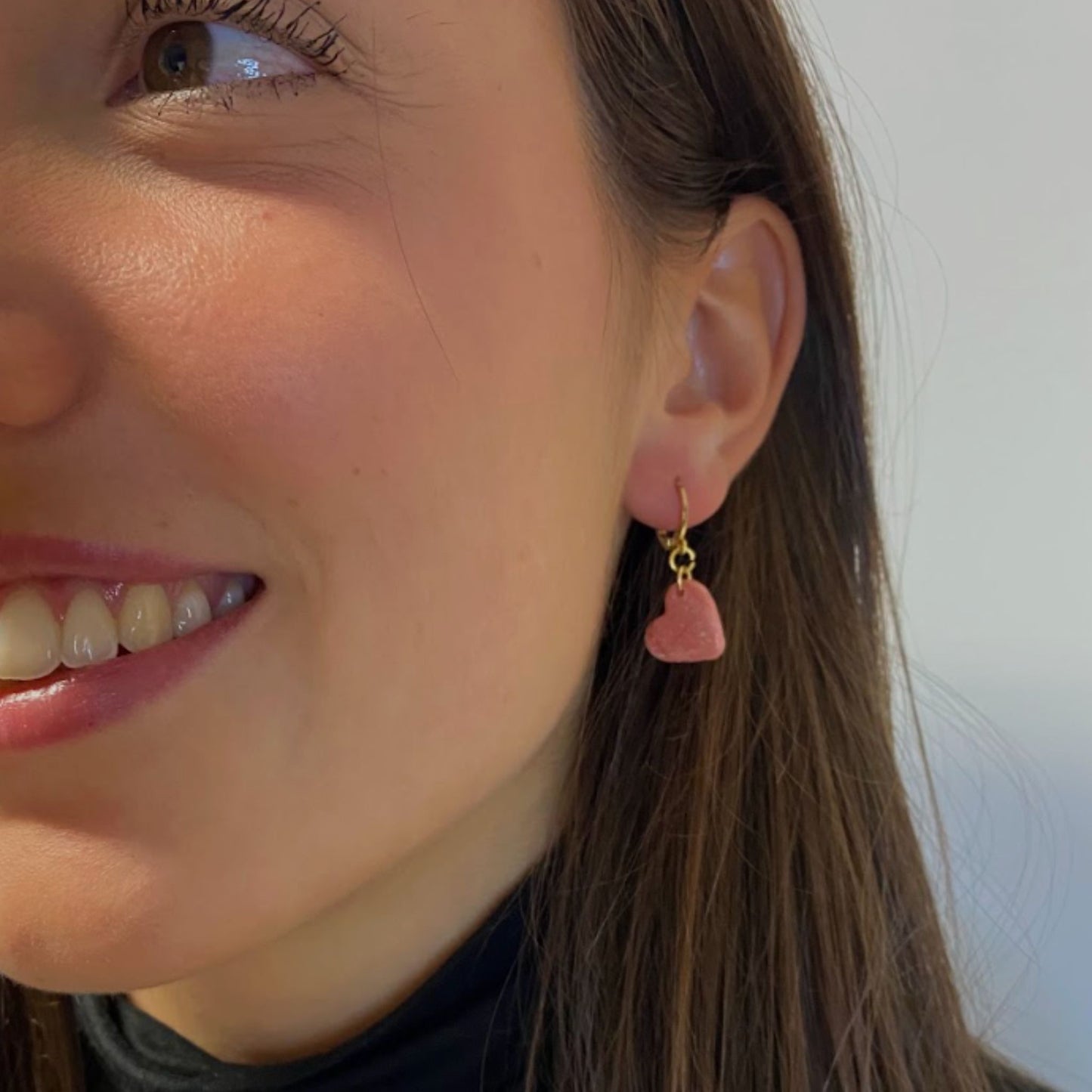 Primo piano di una persona con parte del viso visibile, si vedono parte della bocca sorridente, l’occhio e la guancia. Indossa  un orecchino pendente handmade in porcellana fredda a forma di cuore, colore rosa con venature bianche e dettagli in oro, effetto marmo, con gancio a cerchio a leva, dorato. 
Gioielli fatti a mano da artigiani ciechi e ipovedenti.