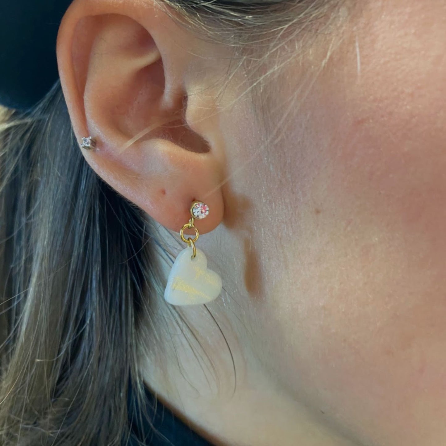 Primo piano di un orecchio che indossa un orecchino pendente handmade in porcellana fredda a forma di cuore, colore bianco con venature dorate. 
L’orecchino ha una chiusura a perno dorata, con punto luce in zircone. 
Orecchini fatti a mano da artigiani ciechi e ipovedenti.
