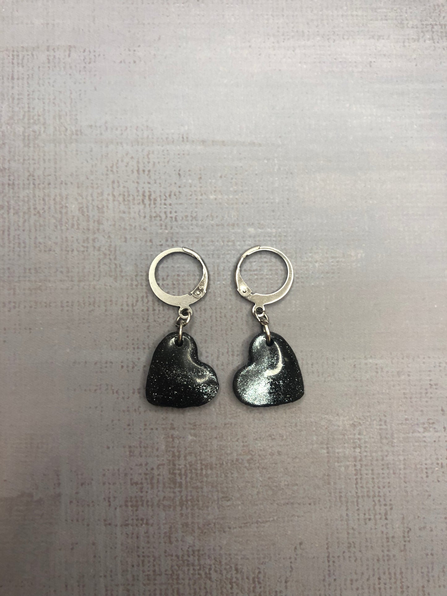Orecchini pendenti artigianali in porcellana fredda a forma di cuore, colore nero con sfumature argento, con gancio a cerchio a leva argentato. 
Gioiello realizzato a mano da artigiani ciechi e ipovedenti.