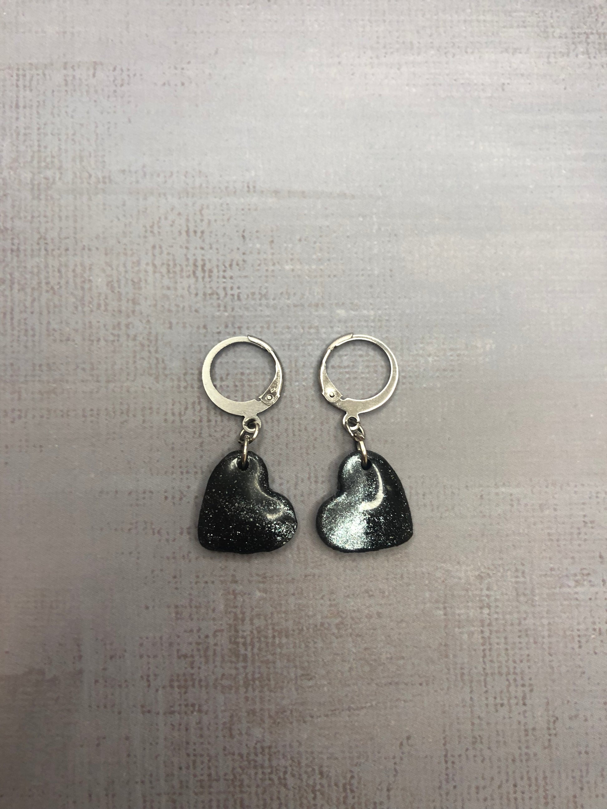 Orecchini pendenti artigianali in porcellana fredda a forma di cuore, colore nero con sfumature argento, con gancio a cerchio a leva argentato. 
Gioiello realizzato a mano da artigiani ciechi e ipovedenti.