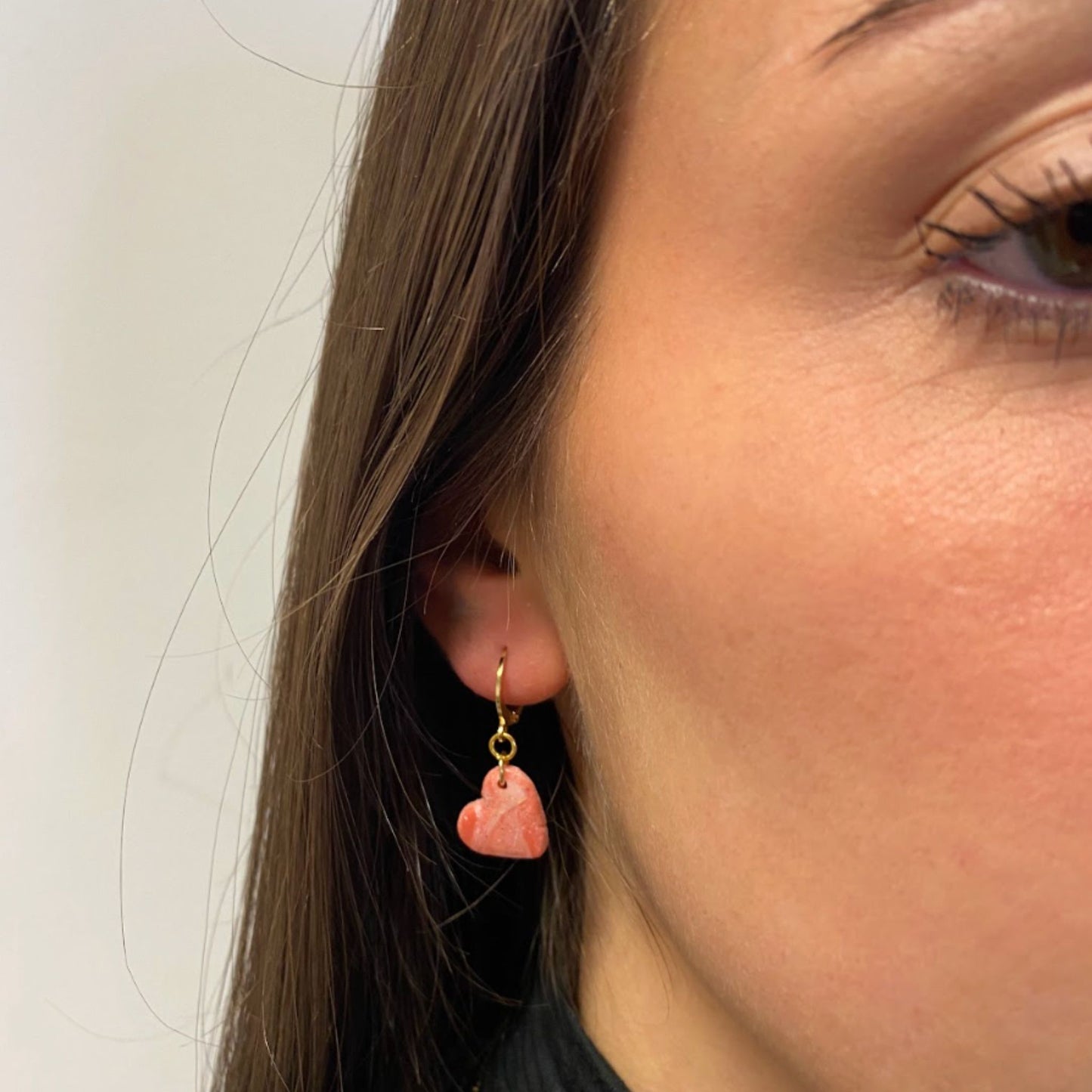Primo piano di una persona con parte del viso visibile, si vedono la guancia e parte dell’occhio. Indossa un orecchino pendente artigianale in porcellana fredda a forma di cuore, colore rosa con venature bianche e dettagli in oro, effetto marmo, con gancio a cerchio a leva, dorato. Gioiello realizzato a mano da artigiani ciechi e ipovedenti. 
