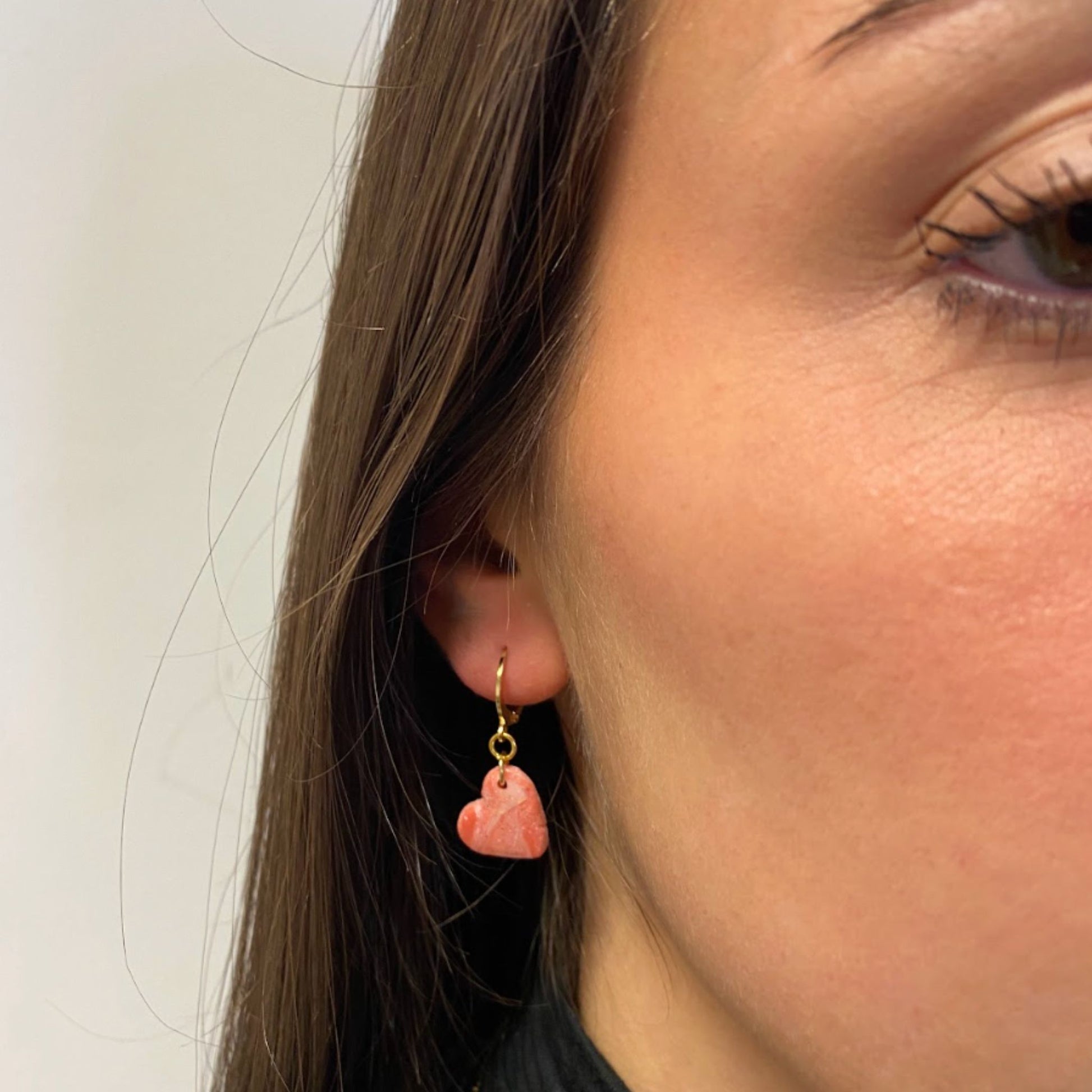 Primo piano di una persona con parte del viso visibile, si vedono la guancia e parte dell’occhio. Indossa un orecchino pendente artigianale in porcellana fredda a forma di cuore, colore rosa con venature bianche e dettagli in oro, effetto marmo, con gancio a cerchio a leva, dorato. Gioiello realizzato a mano da artigiani ciechi e ipovedenti. 
