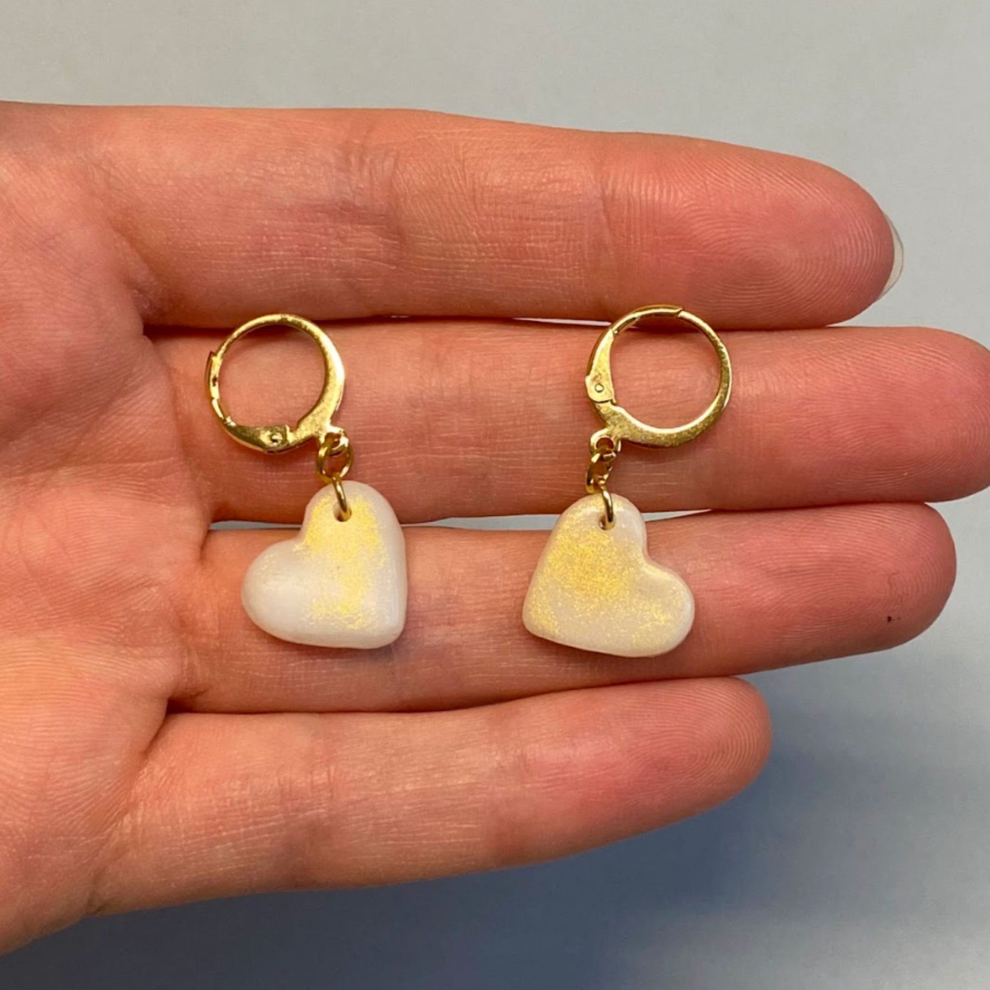 Orecchini pendenti handmade in porcellana fredda a forma di cuore, colore bianco con venature oro, con gancio a cerchio a leva, dorato. 
Fatti a mano da artigiani ciechi e ipovedenti.
Gli orecchini vengono tenuti in mano. 