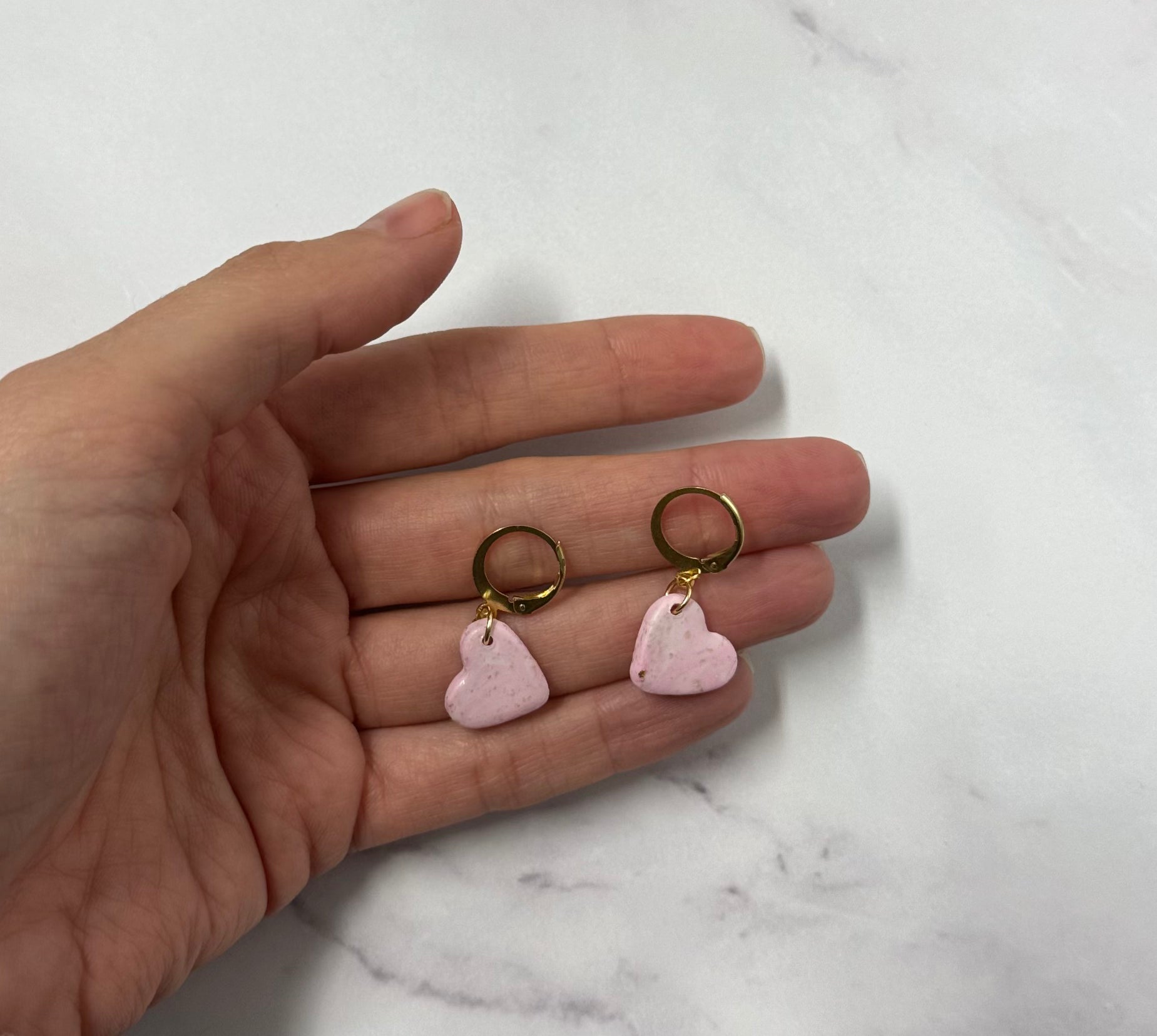 Orecchini con cerchi dorati e ciondolo a forma di cuore rosa in porcellana fredda, sul palmo di una mano. Gioiello realizzato da artigiani ciechi e ipovedenti