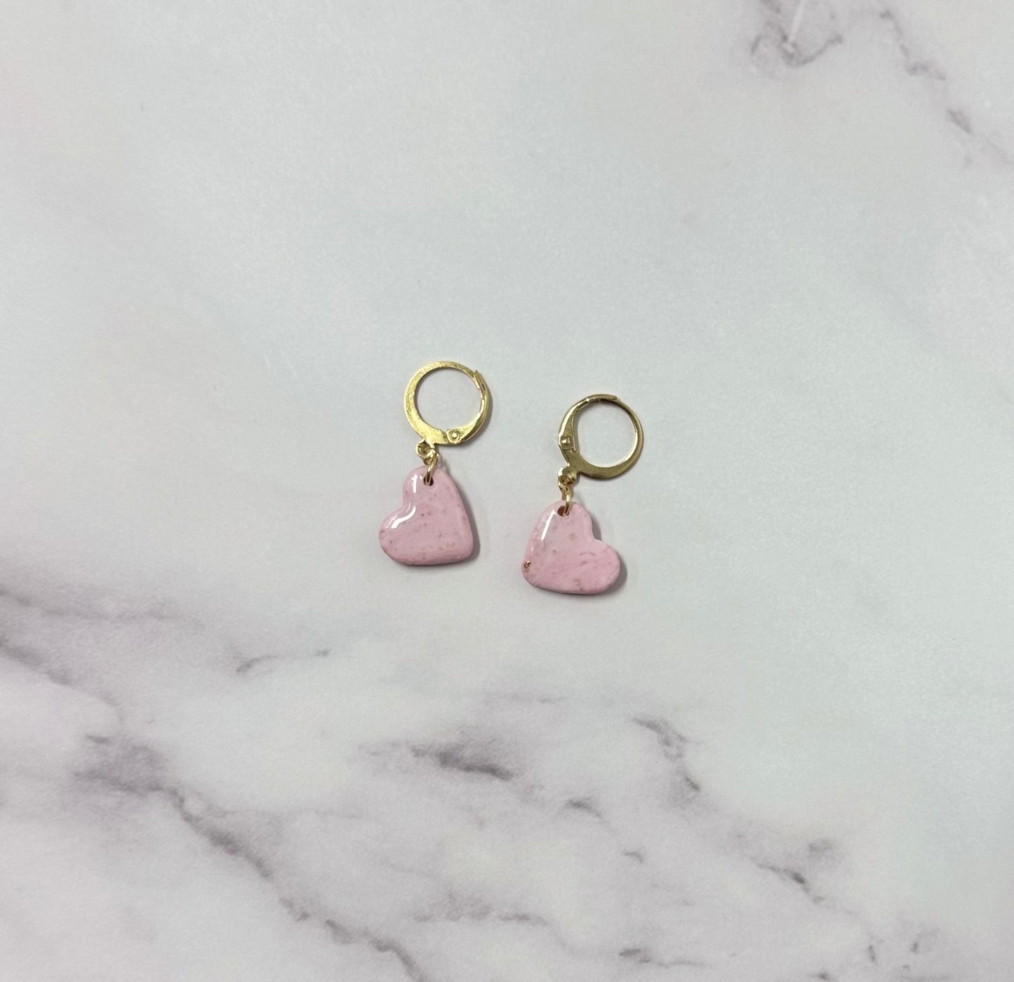 Orecchini con cerchio dorato e ciondolo a forma di cuore rosa in porcellana fredda, adagiata su fondo marmoreo. Creati da artigiani ciechi e ipovedenti.