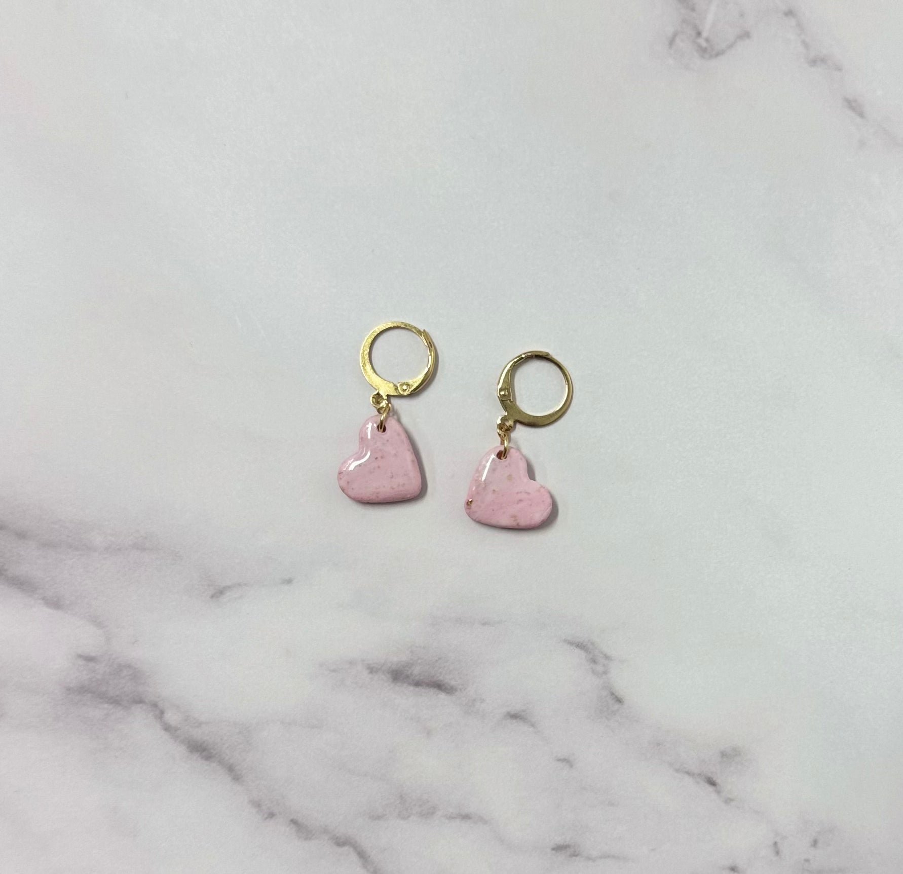 Orecchini con cerchio dorato e ciondolo a forma di cuore rosa in porcellana fredda, adagiata su fondo marmoreo. Creati da artigiani ciechi e ipovedenti.