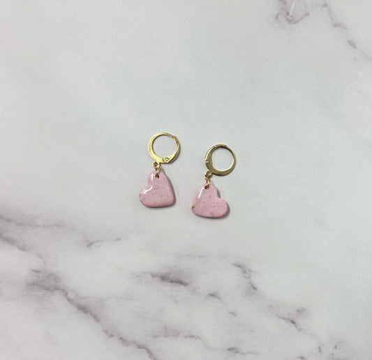 Orecchini con cerchio dorato e ciondolo a forma di cuore rosa in porcellana fredda, adagiata su fondo marmoreo. Creati da artigiani ciechi e ipovedenti.