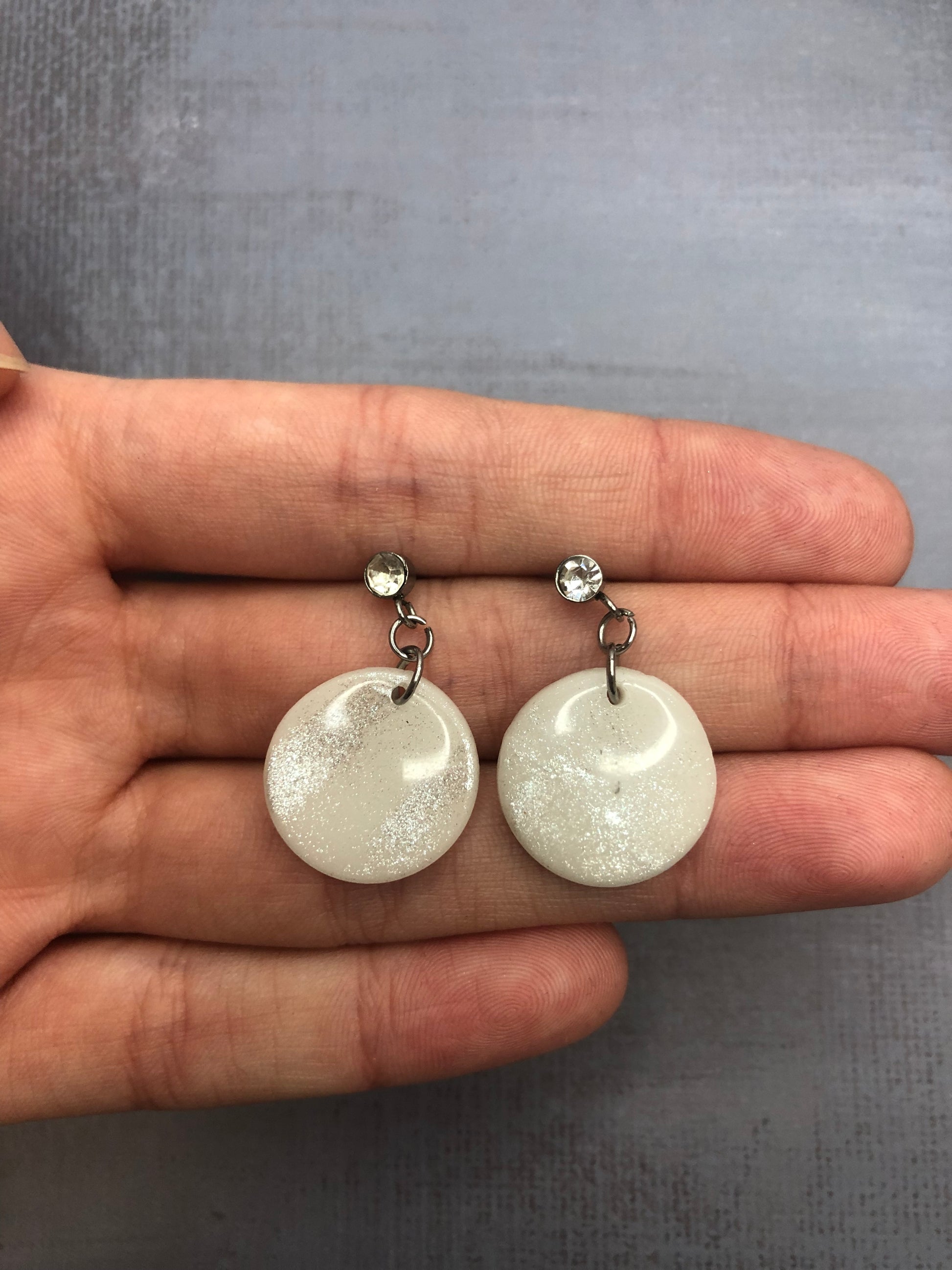 Orecchini pendenti artigianali in porcellana fredda a forma di cerchio, colore bianco con sfumature argento, con chiusura a perno argentata e punto luce in zircone. 
Gioiello realizzato a mano da artigiani ciechi e ipovedenti.
Gli orecchini vengono tenuti in mano. 