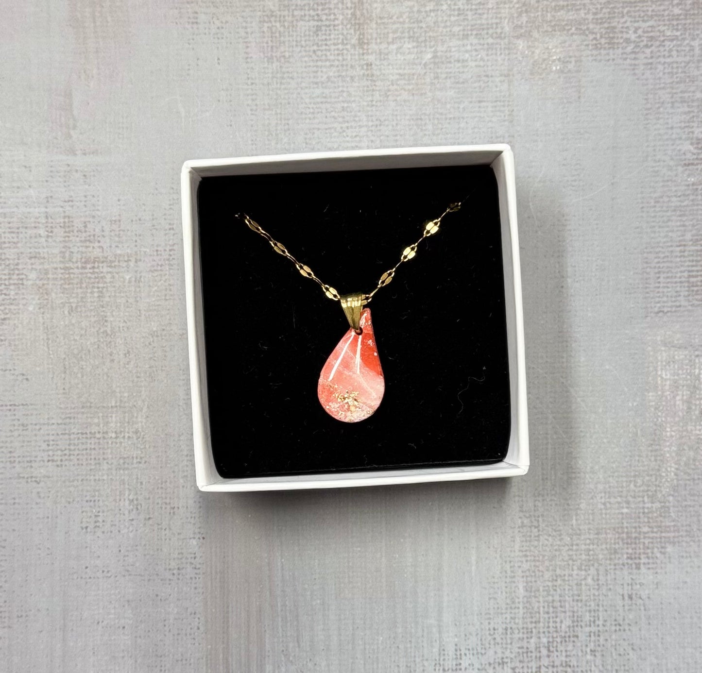 Confezione originale Myra Lab aperta, contenente la collana dorata con ciondolo a forma di goccia rosa marmorizzato, con leggere sfumature dorate, in porcellana fredda. Gioiello artigianale realizzato da persone con disabilità visiva.