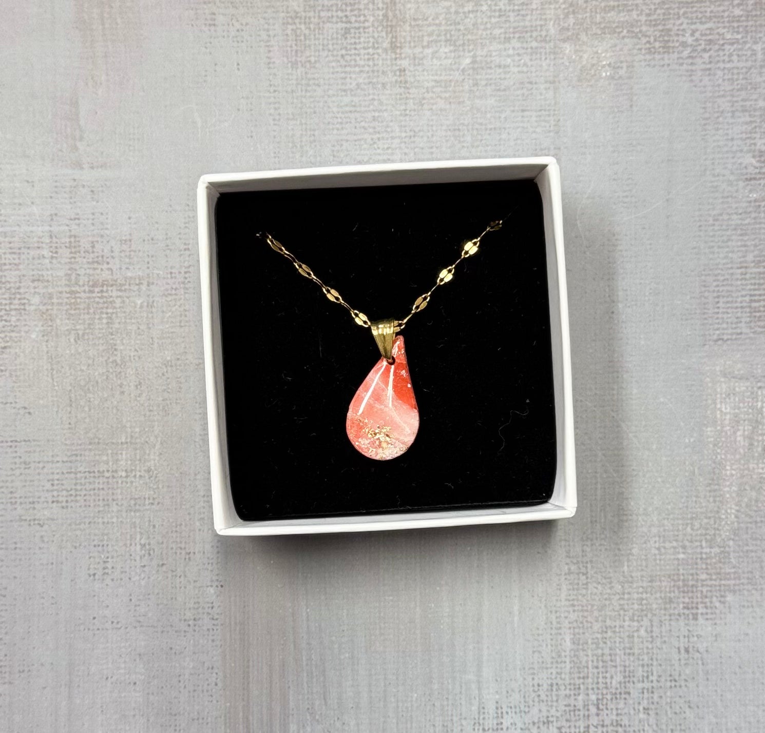 Confezione originale Myra Lab aperta, contenente la collana dorata con ciondolo a forma di goccia rosa marmorizzato, con leggere sfumature dorate, in porcellana fredda. Gioiello artigianale realizzato da persone con disabilità visiva.