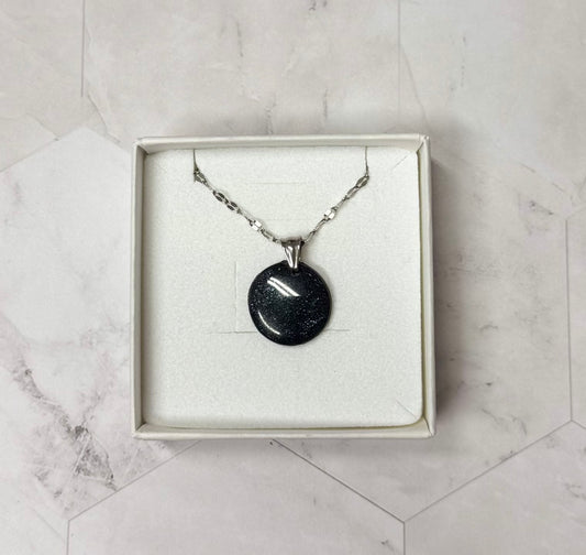 Confezione originale Myra Lab aperta, contenente la collana con pendente tondo nero brillantinato, in porcellana fredda. Gioiello artigianale realizzato da persone cieche e ipovedenti.