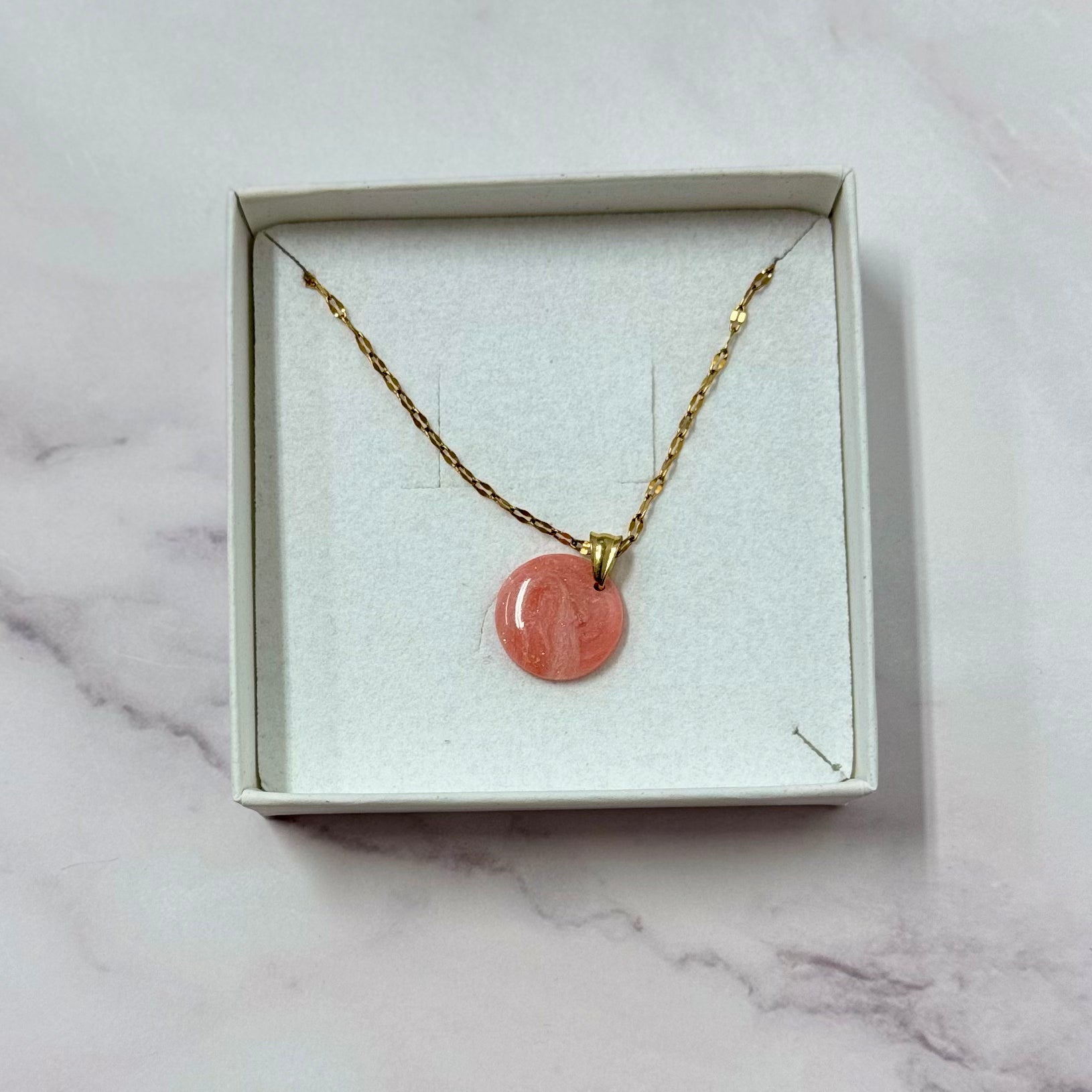 Confezione originale Myra Lab aperta, contenente la collana con ciondolo tondo rosso/rosa marmorizzato in porcellana fredda con leggere sfumature dorate. Gioiello artigianale realizzato da persone cieche e ipovedenti.