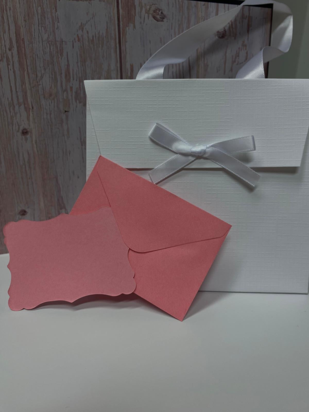 Confezione regalo bianca con fiocco in raso, affiancata da bigliettino e busta rosa.